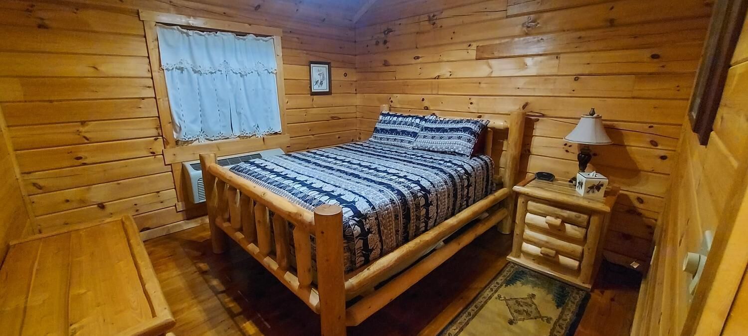 Una camera in una baita di legno, arredata con un letto, un comodino, una lampada e una finestra con tende.