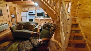 Un interior de cabaña acogedor con sofá y sillón, una escalera y una zona de cocina.