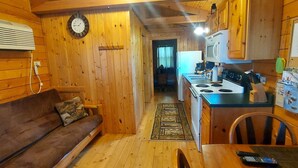 A compact wooden cabin with a kitchen area featuring a stove, microwave, and refrigerator. A wooden bench and a small table with a remote control are visible. - Walleye -  Sleeps up to Five (Moravia)