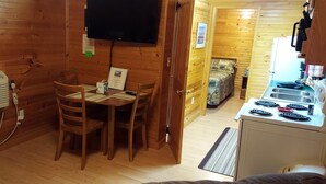 A wooden cabin interior with a dining area, a kitchen with a stove, and a television mounted on the wall.