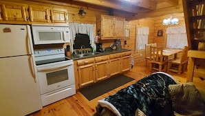 A wooden cabin interior with a kitchen area featuring a refrigerator, microwave, oven, and wooden cabinets. There is a dining table with chairs and a dog bed in the living area. - Elk - Sleeps up to 6 (Moravia)