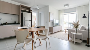 A modern kitchen with a dining area, a sofa, and a TV in a living room. - Cannes Club by Viva - Residence a 200m da praia | Ingleses do Rio Vermelho (Florianópolis)