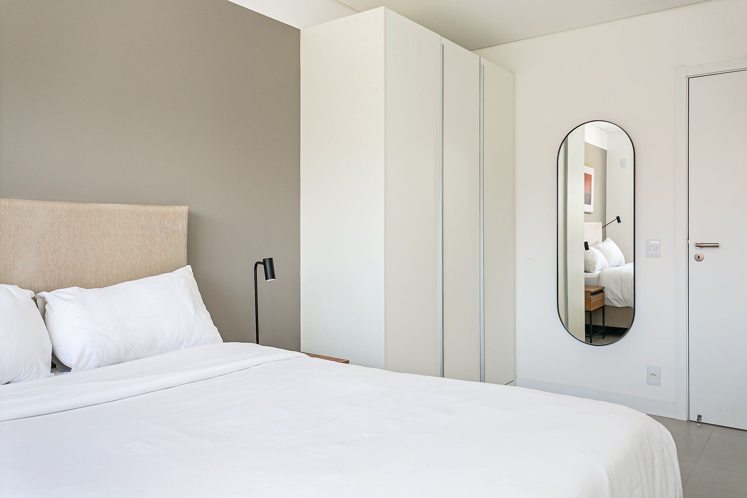 A neatly made bed with white linens, a bedside lamp, and a large mirror on the wall.