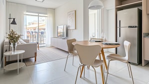 A modern living room with a sofa, a dining table with chairs, a kitchen area with a refrigerator, and a large window with curtains. - Cannes Club by Viva - Residence a 200m da praia | Ingleses do Rio Vermelho (Florianópolis)