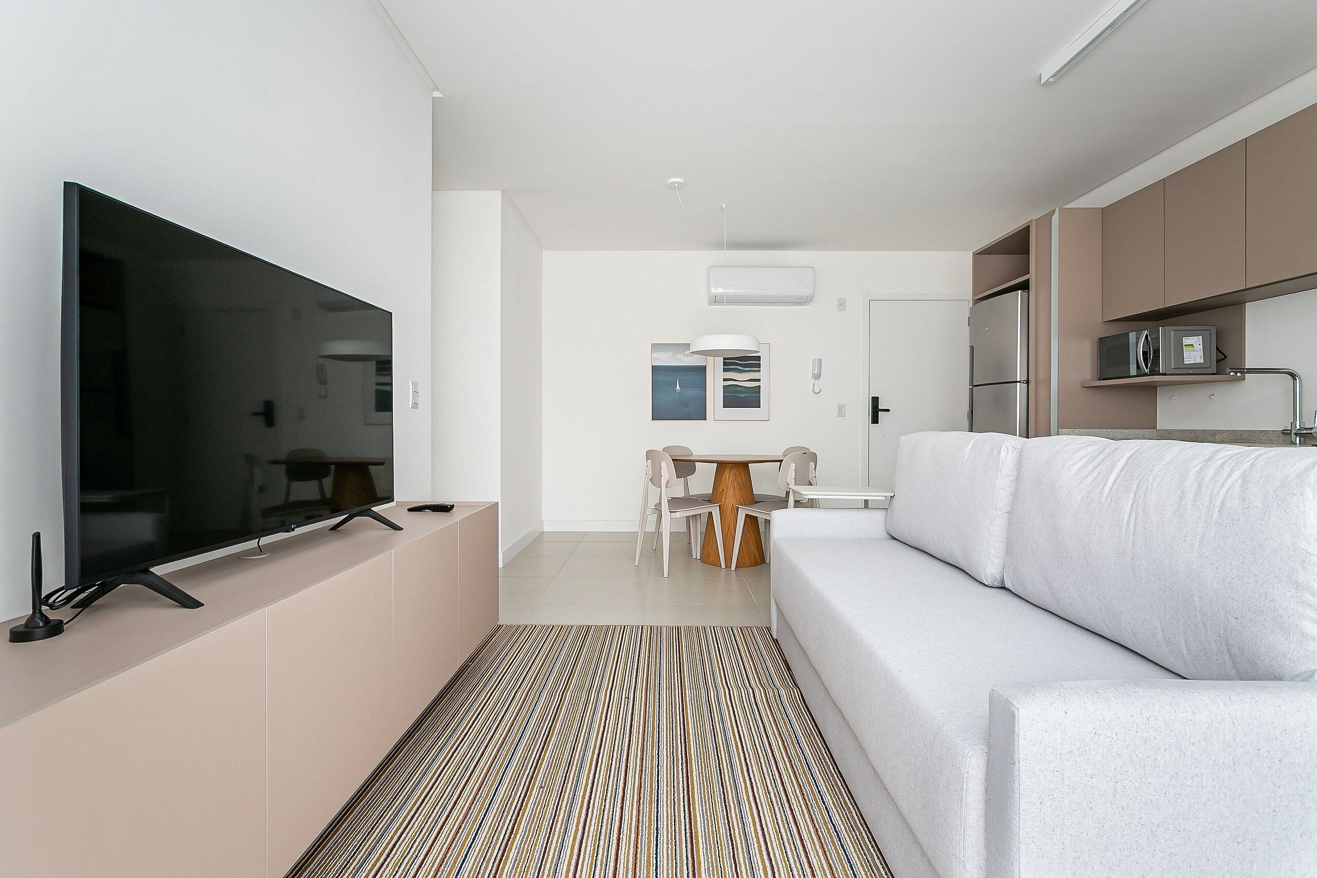 Uma sala de estar moderna com televisão de tela plana, sofá branco, mesa de jantar com cadeiras e área de cozinha com armários e eletrodomésticos.