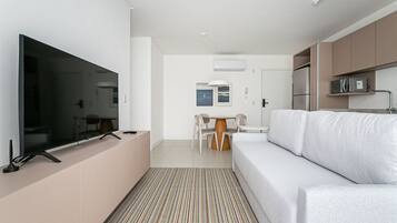 A modern living room with a flat-screen TV, a white sofa, a dining table with chairs, and a kitchen area with cabinets and appliances.