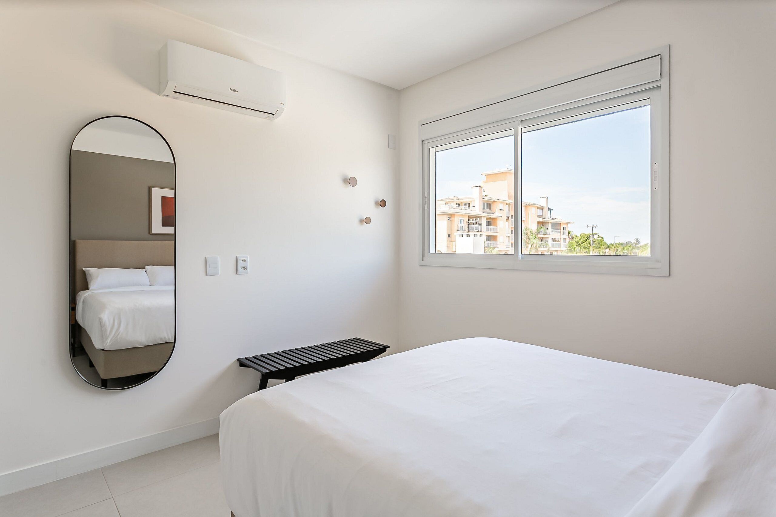 A modern hotel room with a large bed, a bedside table, a mirror, and a window with a view of buildings.