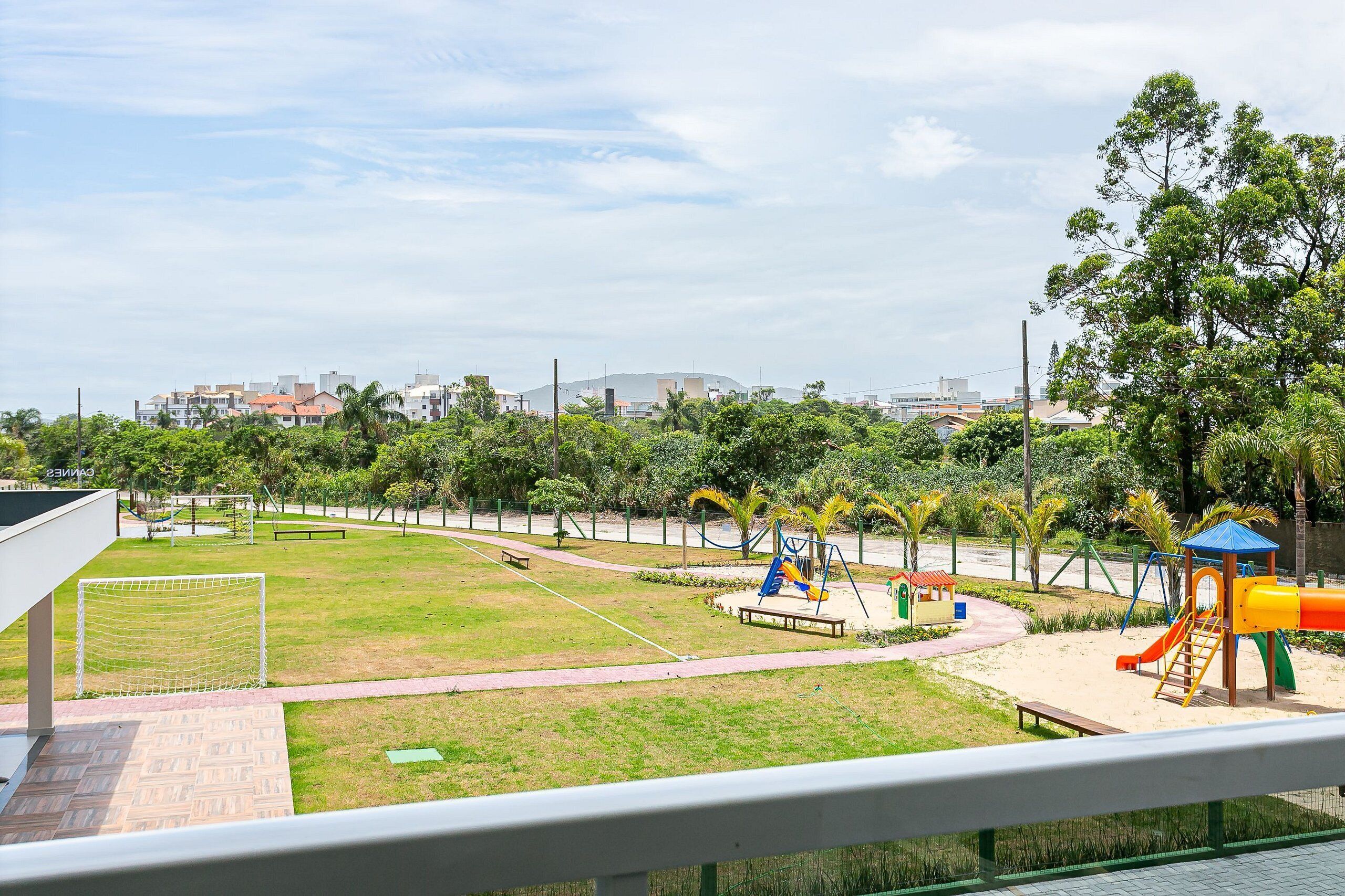 A playground with a soccer goal, swings, and slides, surrounded by greenery and buildings in the background.