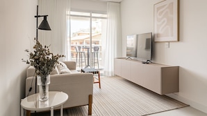 A modern living room with a sofa, a small table with a vase, a TV on a cabinet, and a view of the outside through large windows. - Cannes Club by Viva - Residence a 200m da praia | Ingleses do Rio Vermelho (Florianópolis)