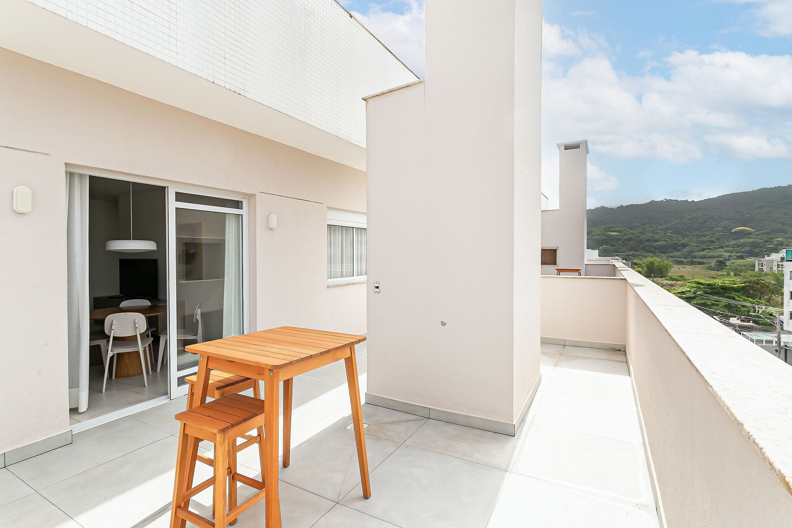 Un balcón con una mesa y sillas de madera, una puerta corredera de vidrio que da a un espacio interior y vistas a un paisaje verde.