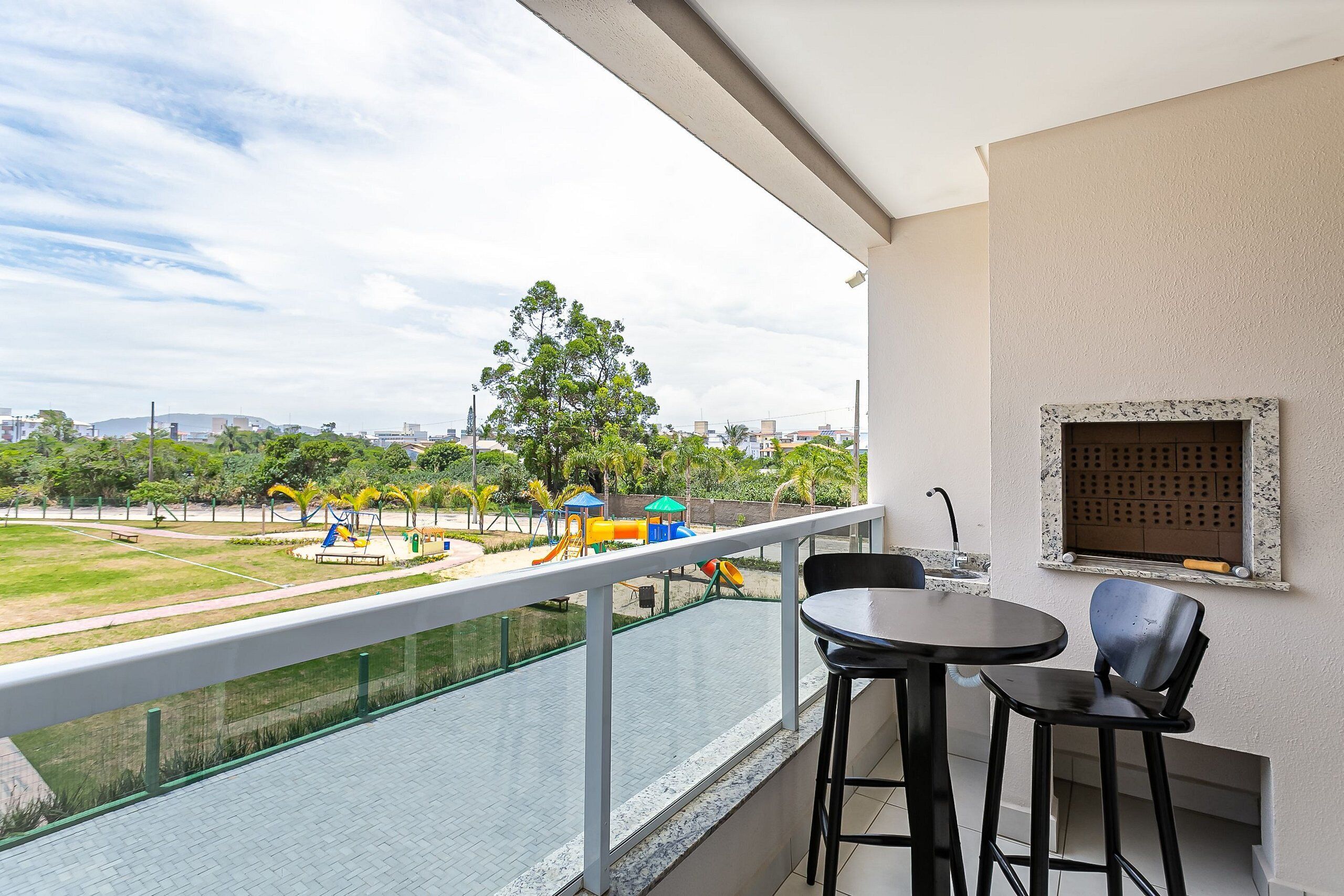 Un balcón con vista a un parque infantil, una mesa con sillas y un ventanal con marco decorativo.
