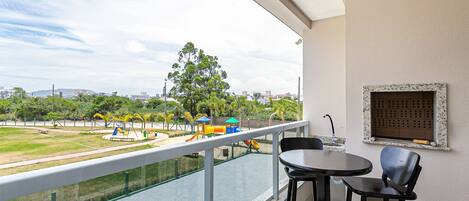 Un balcón con vista a un parque infantil, una mesa con sillas y un ventanal con marco decorativo.