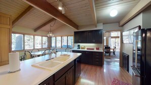 A spacious kitchen with wooden beams, a large island with a sink, dark cabinets, and a refrigerator. - Spacious Cabin Vibe w/ lots of Wood - Easy walk/ride to Village, Lodge and River (Sunriver)