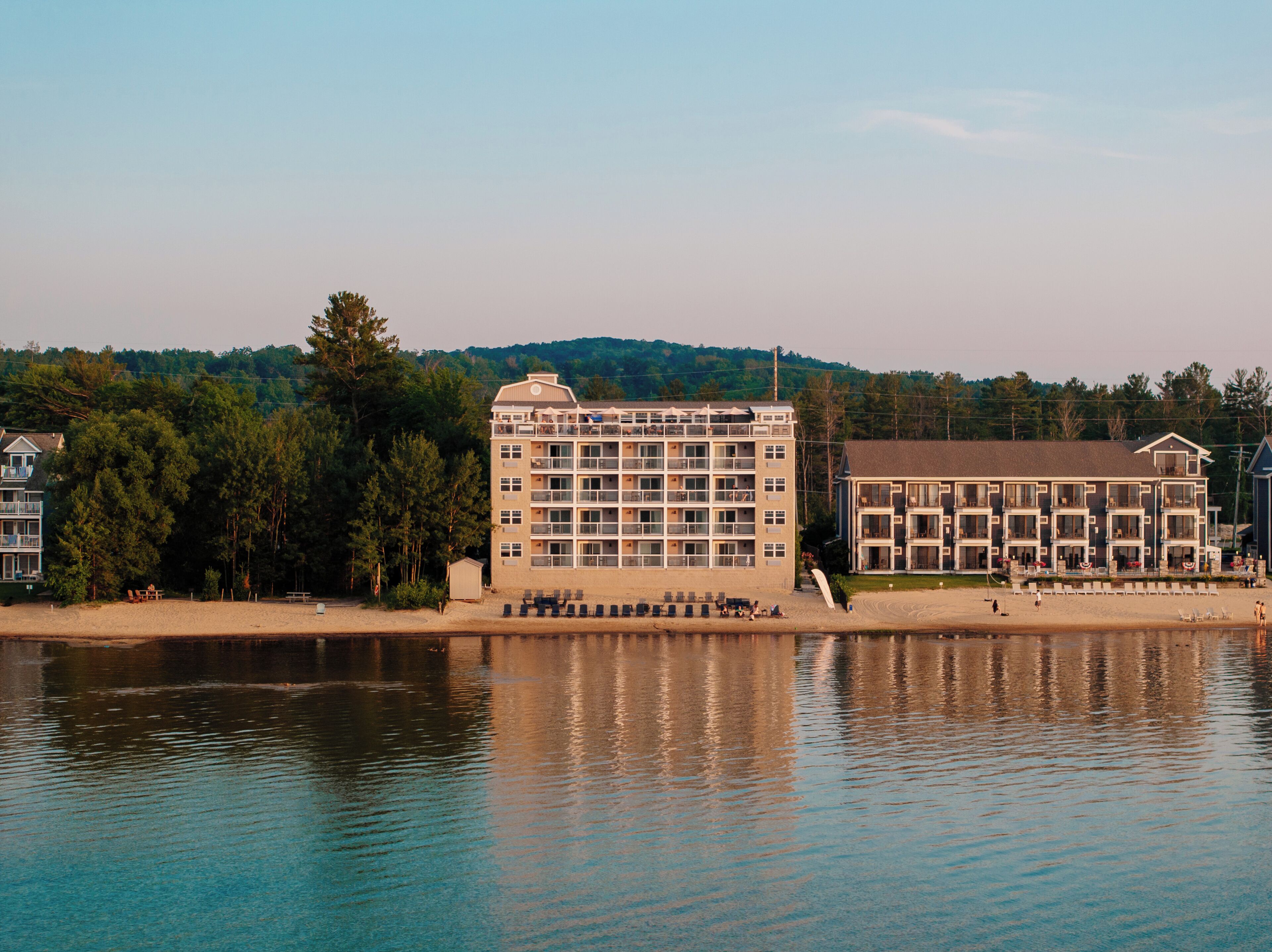 A multi-story hotel with balconies, situated by a lake with a forested hillside in the background.