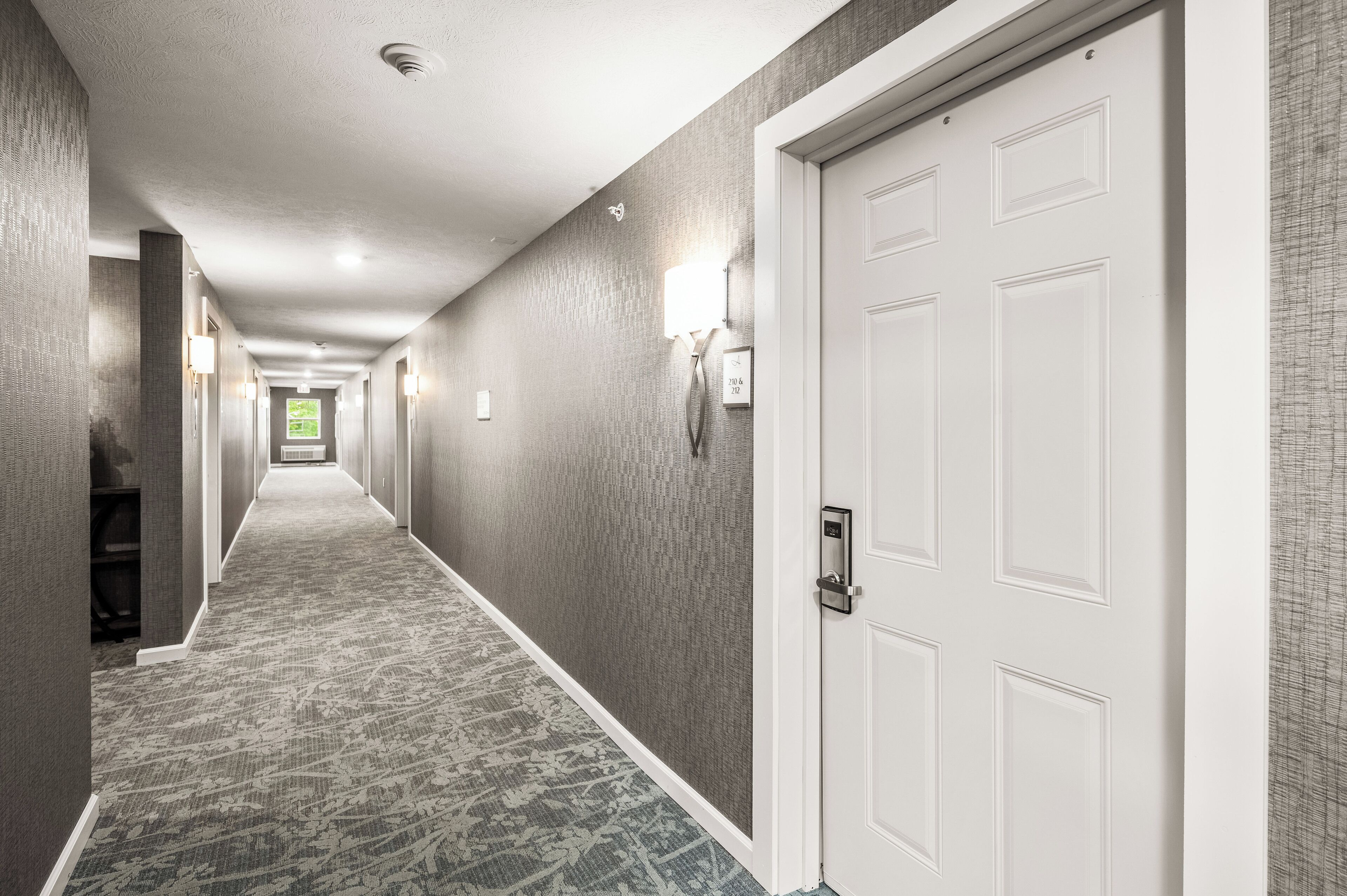 A hotel corridor with patterned carpet, white doors, and wall-mounted light fixtures.