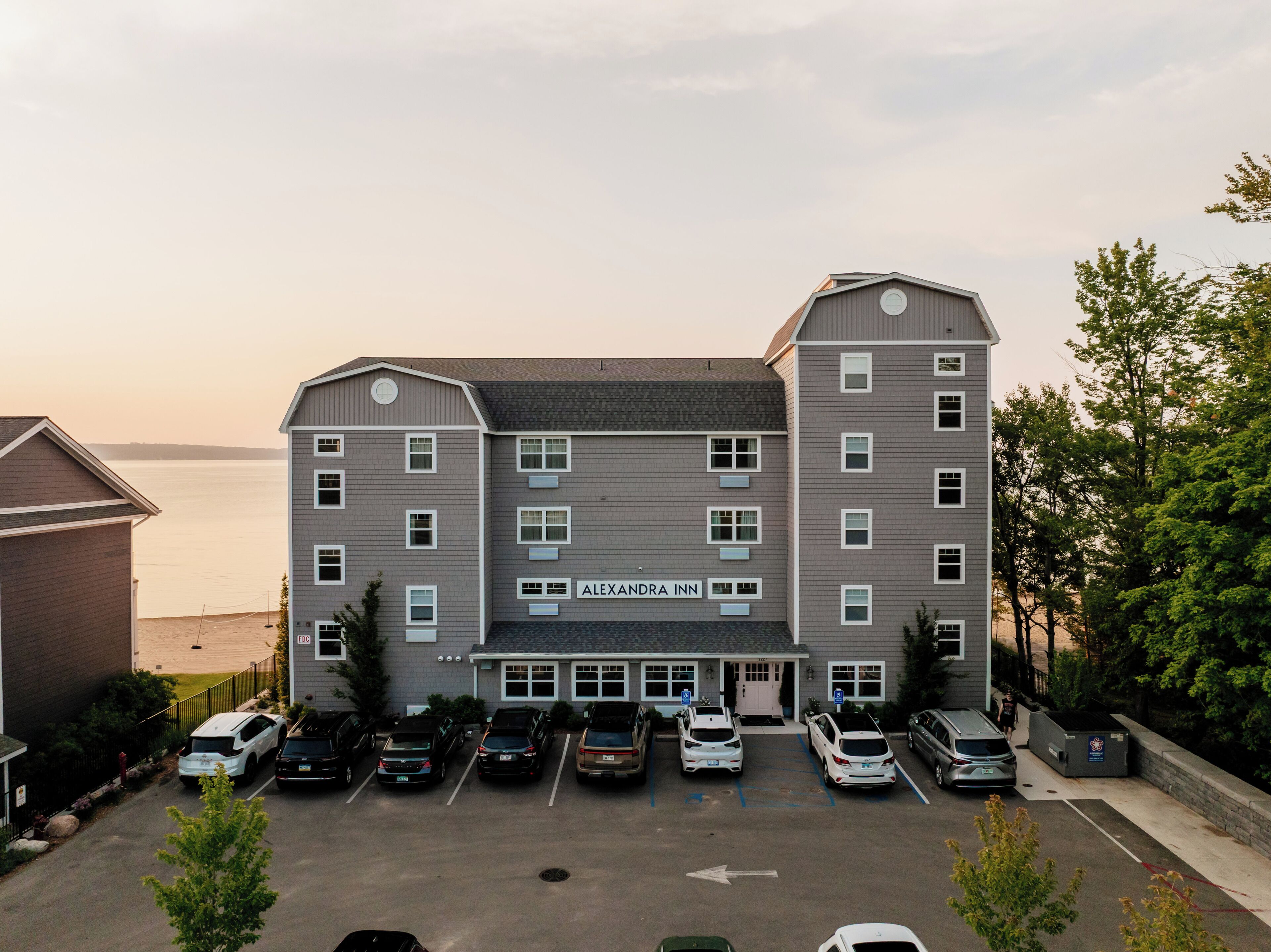 A multi-story hotel with a parking lot in front, a sign reading 'ALEXANDRA INN', and a view of the water in the background.