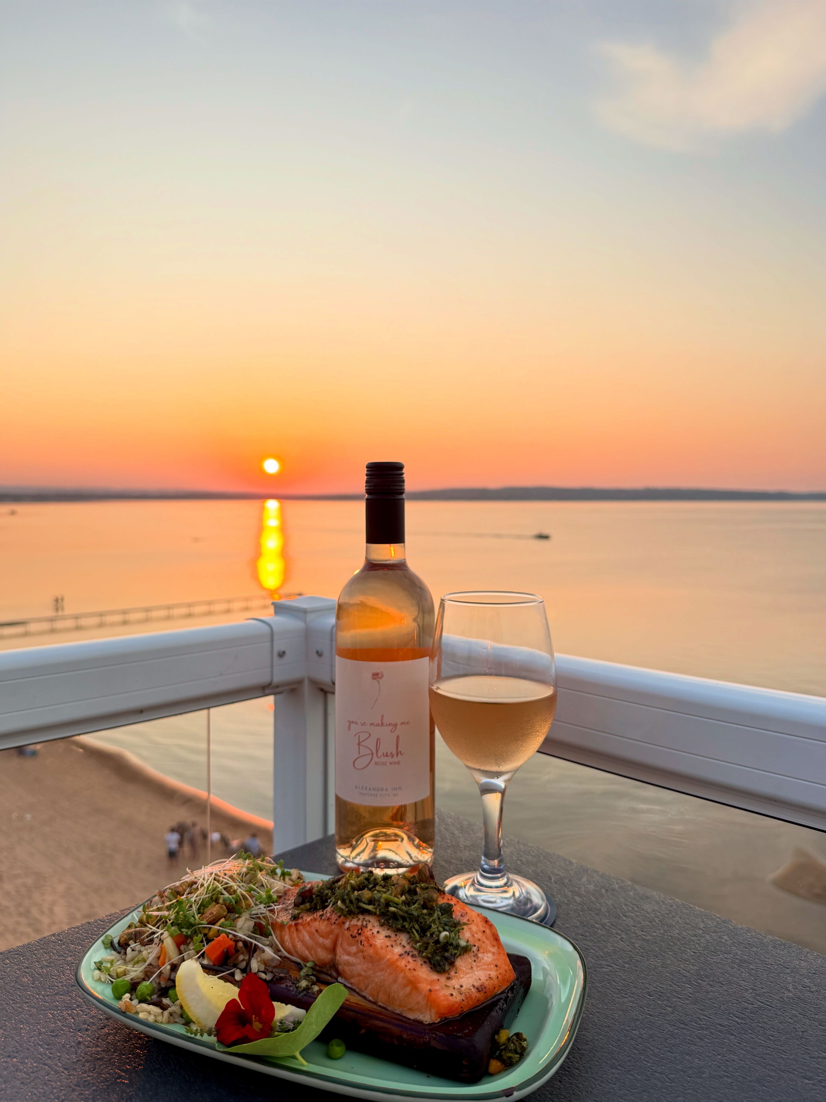 A table with a bottle of wine, a glass of wine, and a plate of food on a balcony overlooking a sunset.