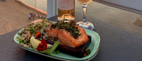 A table with a bottle of wine, a glass of wine, and a plate of food on a balcony overlooking a sunset.
