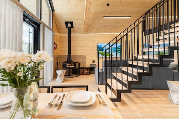 A modern interior with a wooden floor, a fireplace, a staircase with black railings, and a dining area with a vase of flowers.