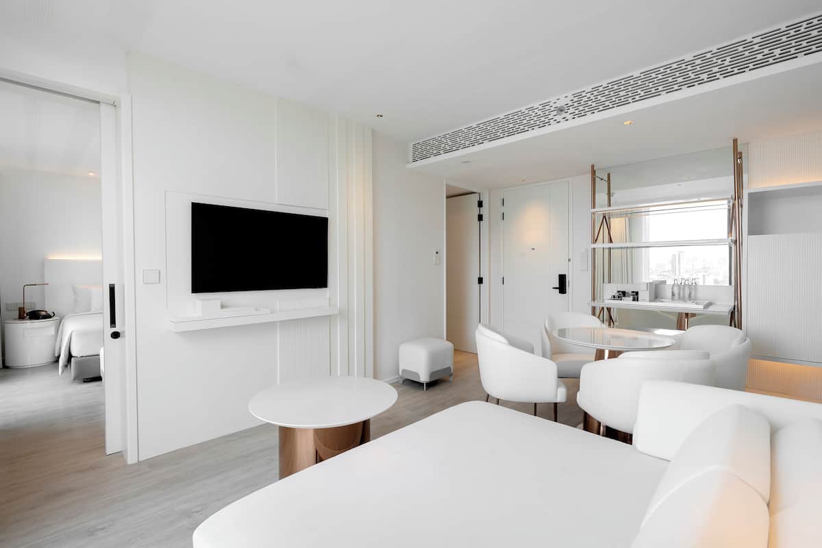A modern hotel room with a flat-screen TV mounted on the wall, a dining area with a round table and white chairs, and a seating area with a white sofa and ottoman.