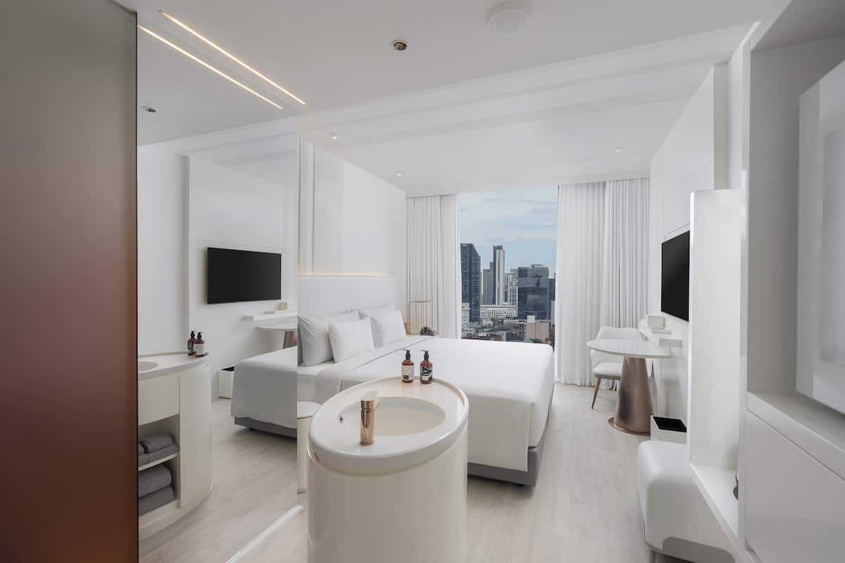 A modern hotel room with a large bed, a bedside table, a round sink, and a city view.