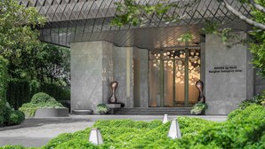 A modern hotel entrance with a geometric facade, a glass door entrance, and a well-maintained garden area.