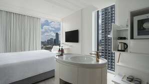 A modern hotel room with a large bed, a central sink, a flat-screen TV, and a city view.