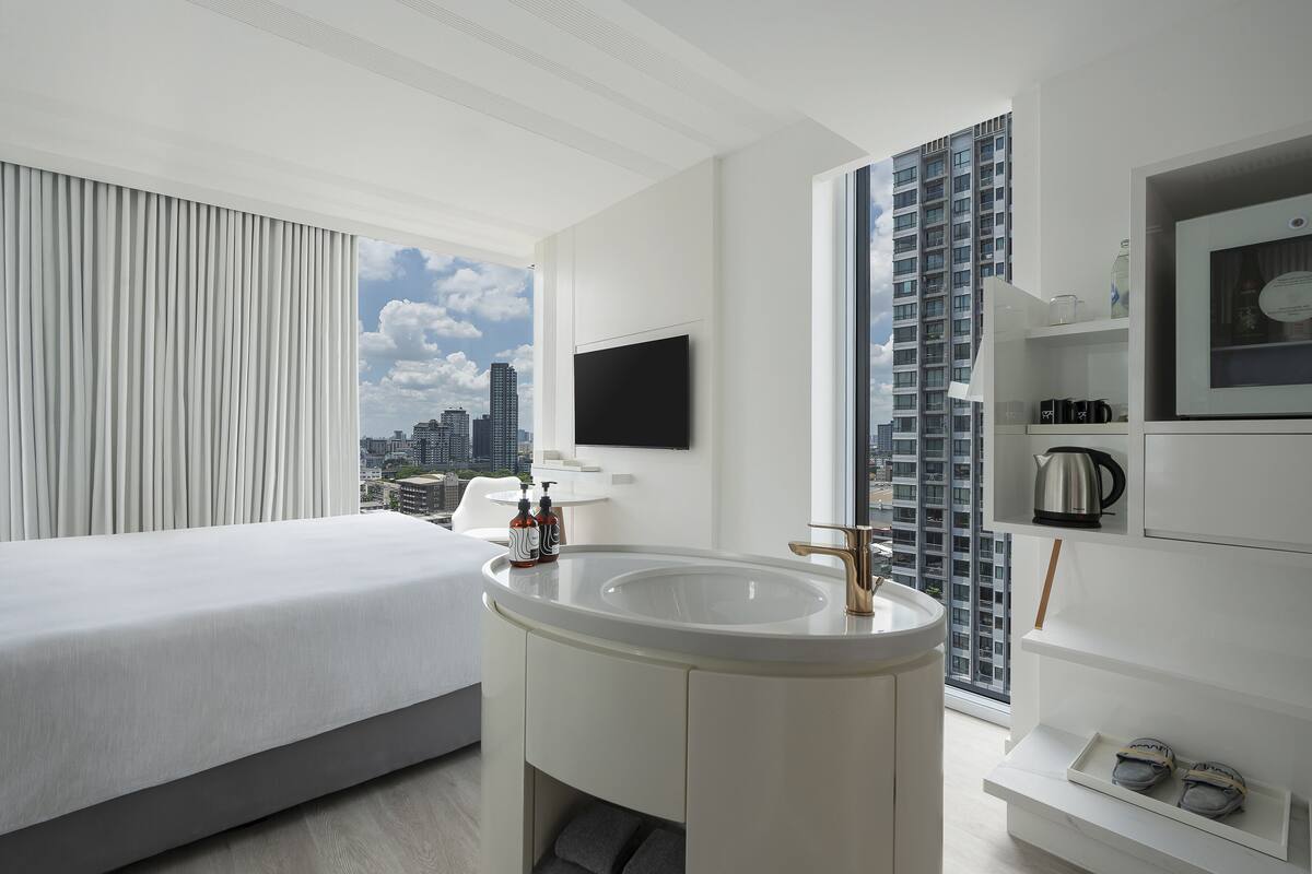 A modern hotel room with a large bed, a central sink, a flat-screen TV, and a city view.