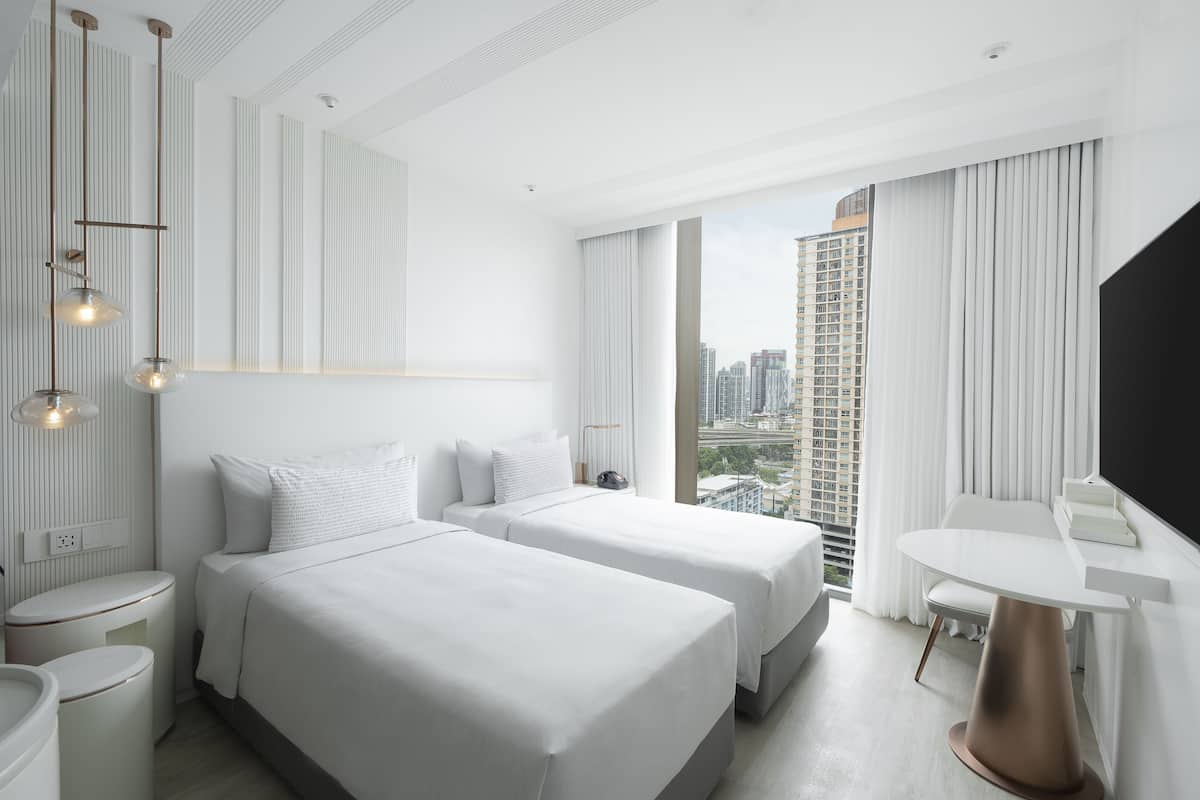 A modern hotel room with two beds, a small table, and a large window offering a city view.