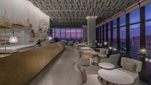 A modern bar area with a high ceiling, large windows, and a city view at dusk.