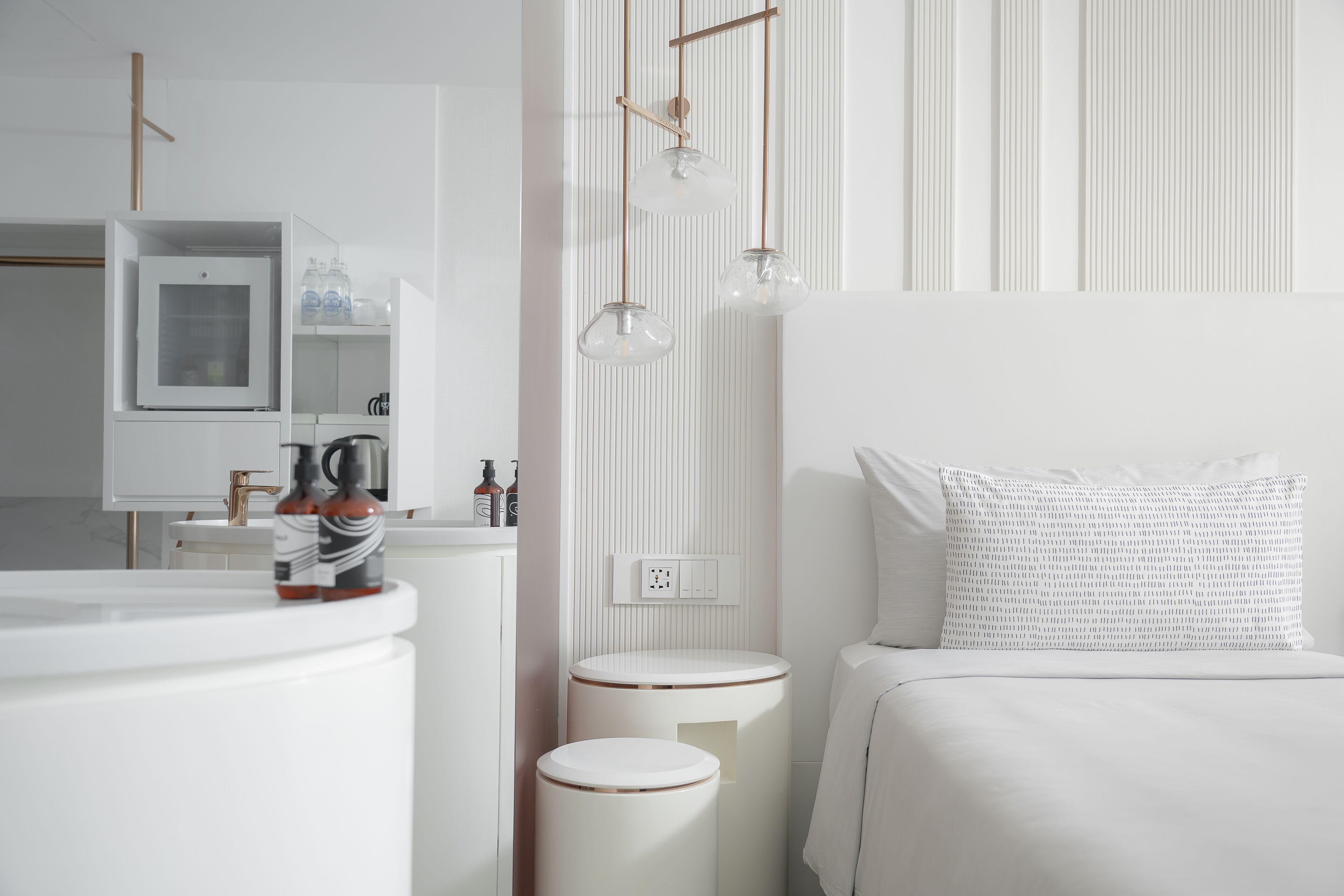 Une chambre d’hôtel moderne aux couleurs claires, avec un lit agrémenté d’un coussin à motifs, une table de chevet sur laquelle se trouve une bouteille, et un miroir au-dessus du lavabo.