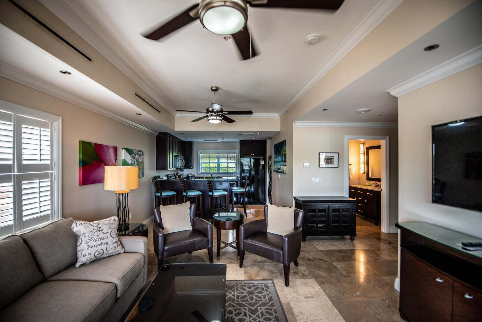 A spacious living room with a sofa, two armchairs, a coffee table, and a TV. A ceiling fan and a kitchen are visible in the background.