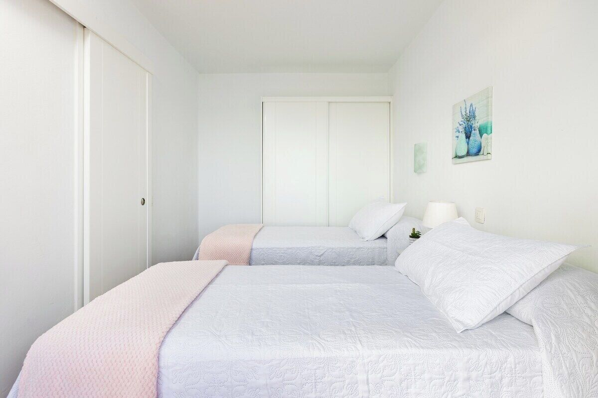 A bedroom with two beds, white bedding, a door, and a wall-mounted lamp.