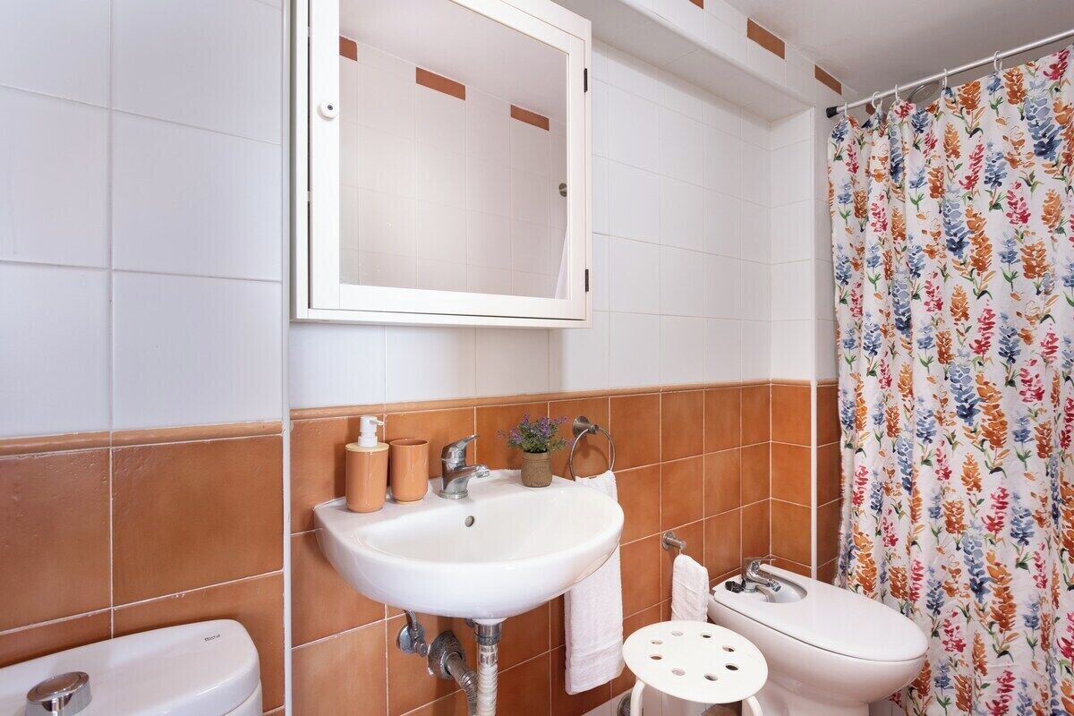 Et badeværelse med en hvid vask, et spejl, et toilet og en bruseforhæng med blomstermønster.