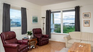 Een woonkamer met twee leren fauteuils, een bank, een salontafel en een vloerlamp. Twee ramen met gordijnen, een lijst met een foto aan de muur, en uitzicht op groen en een brug.