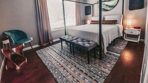 Een slaapkamer met een hemelbed, een nachtkastje met een lamp, een stoel en een kamerbreed tapijt met een patroon.