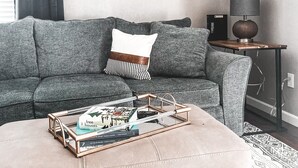 Een moderne woonkamer met een grijze bank, een ronde wandklok, een bijzettafeltje met een lamp, en een salontafel met boeken.