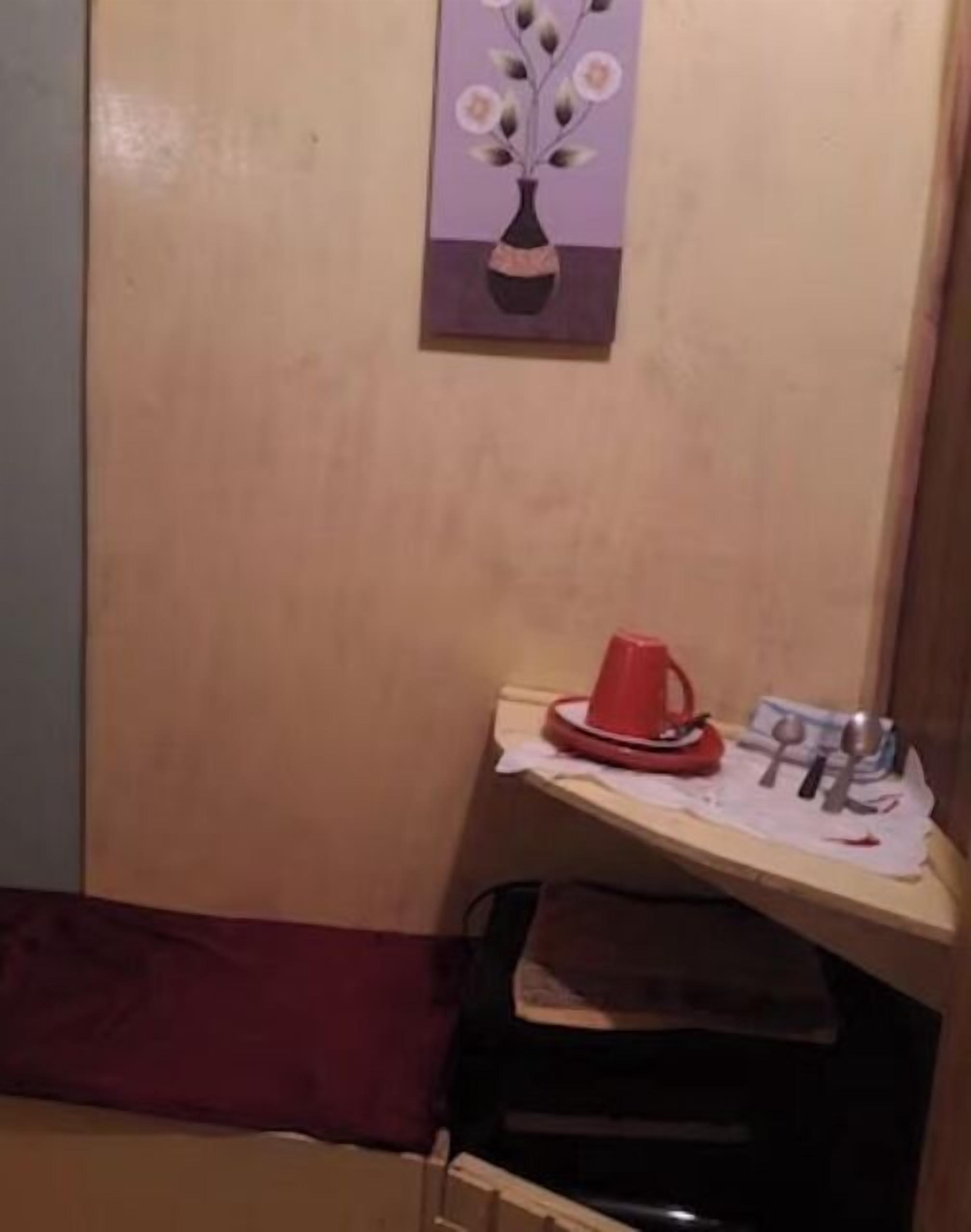 Een klein kitchenette gedeelte met een rode beker en een set lepels op een plank, een vaas met bloemen aan de muur, en een houten bankje.