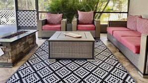 Une terrasse ensoleillée, meublée de mobilier en osier, avec un foyer et un tapis à motifs.