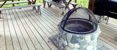 A stone fire pit with a mesh cover on a wooden deck with picnic tables.