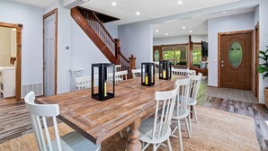 A spacious dining area with a wooden table, white chairs, and black candle holders. A wooden staircase leads to another room, and a large wooden door with a glass panel is visible. - Wine Country Escape - Hot Tub - Sleeps 12 (Geneva)