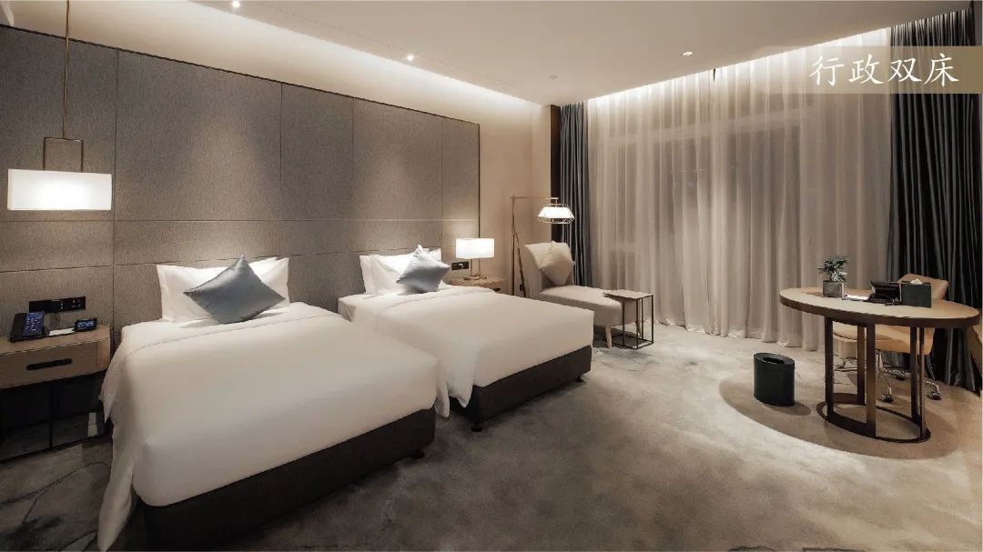 Una habitación de hotel con dos camas, una mesita, una silla, una lámpara y un ventanal con cortinas.