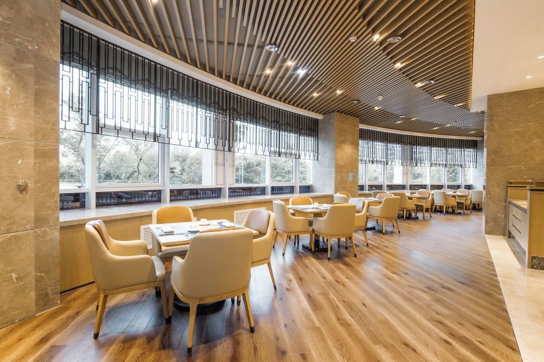 Un espacio interior amplio con piso de madera, varias mesas y sillas, ventanas grandes con vista a árboles y un diseño de techo moderno.