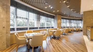 Un espacio interior amplio con piso de madera, varias mesas y sillas, ventanas grandes con vista a árboles y un diseño de techo moderno.