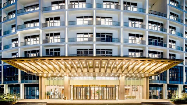 Een hotel met meerdere verdiepingen, met een modern ontwerp, en een opvallende ingang met een glazen gevel en een overdekt gebied met een gouden balustrade.