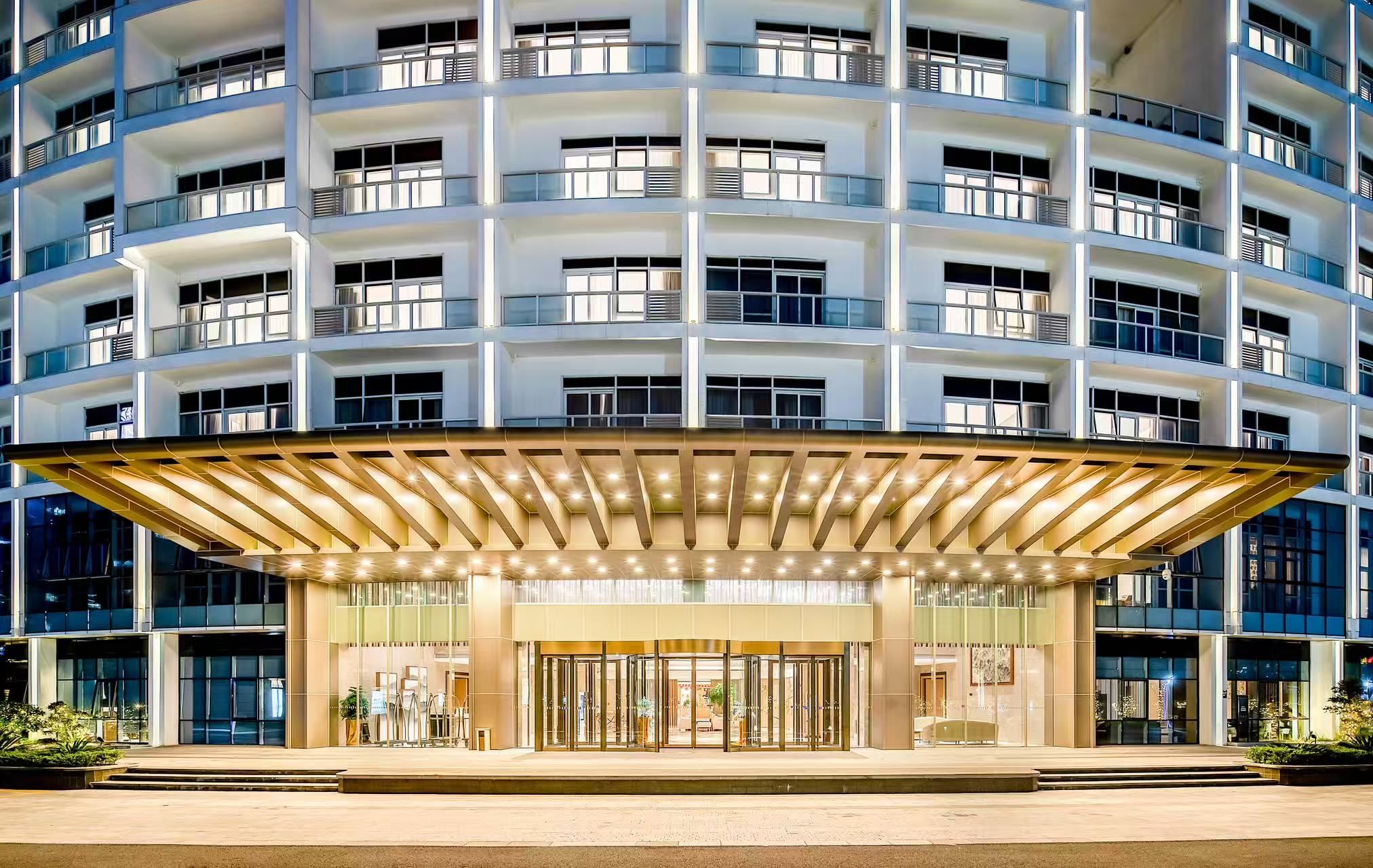 A multi-story hotel with a modern design, featuring a prominent entrance with a glass facade and a covered area with a golden railing.
