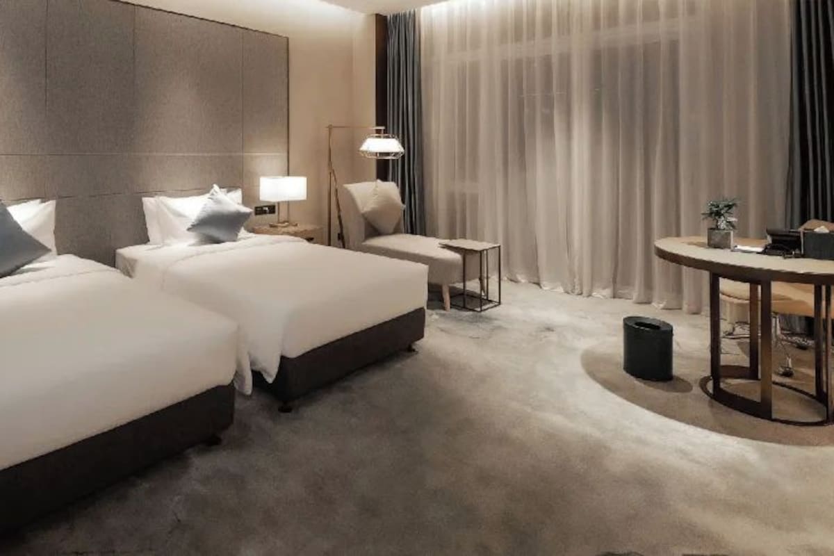 Hotellihuone, jossa on kaksi sänkyä, pieni pöytä, tuoli, lamppu ja suuri ikkuna, jossa on verhot.