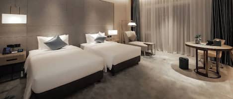 A hotel room with two beds, a small table, a chair, a lamp, and a large window with curtains.