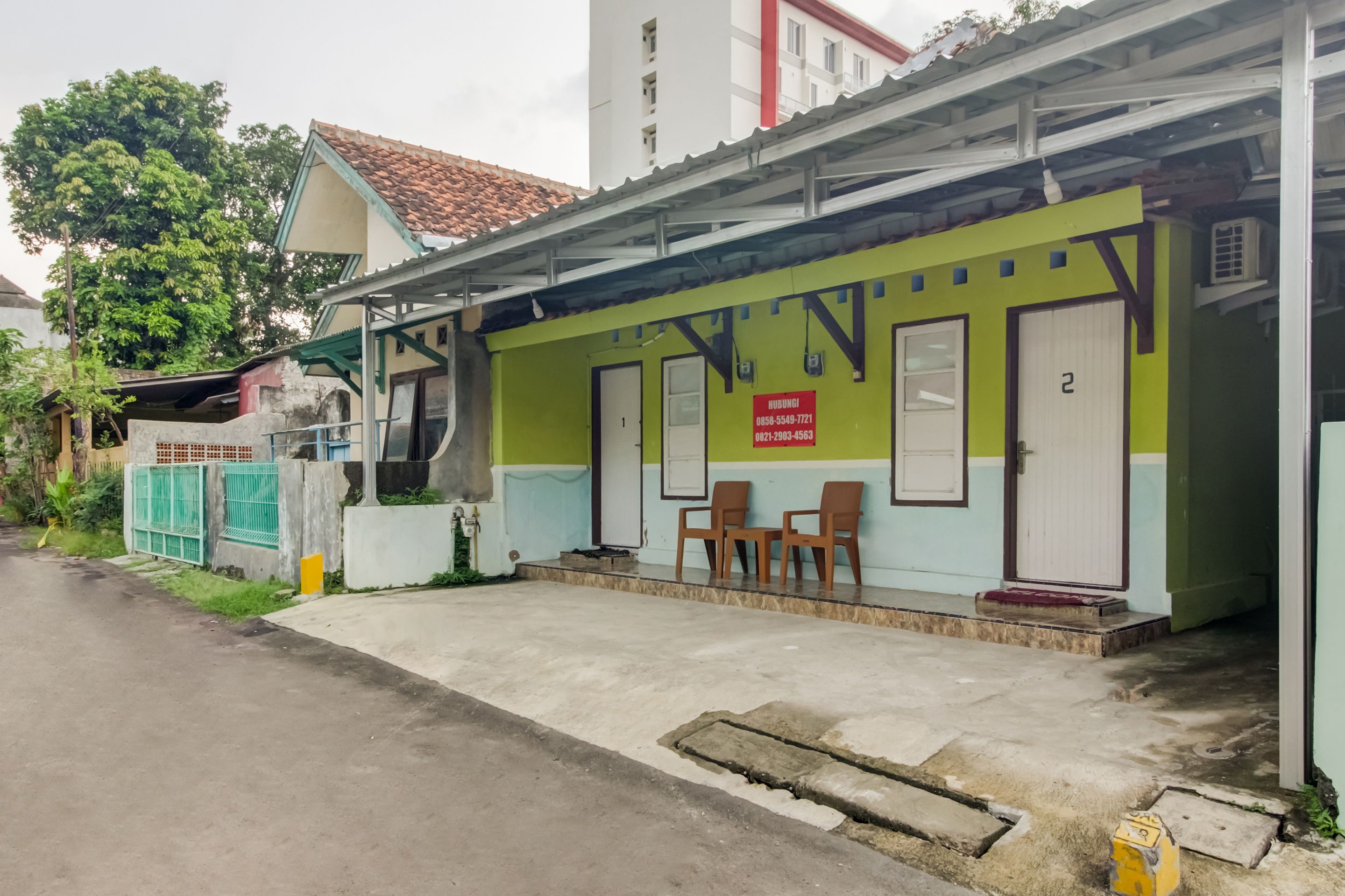 A building with a green and white facade, a red sign with contact numbers, and a set of wooden chairs outside.
