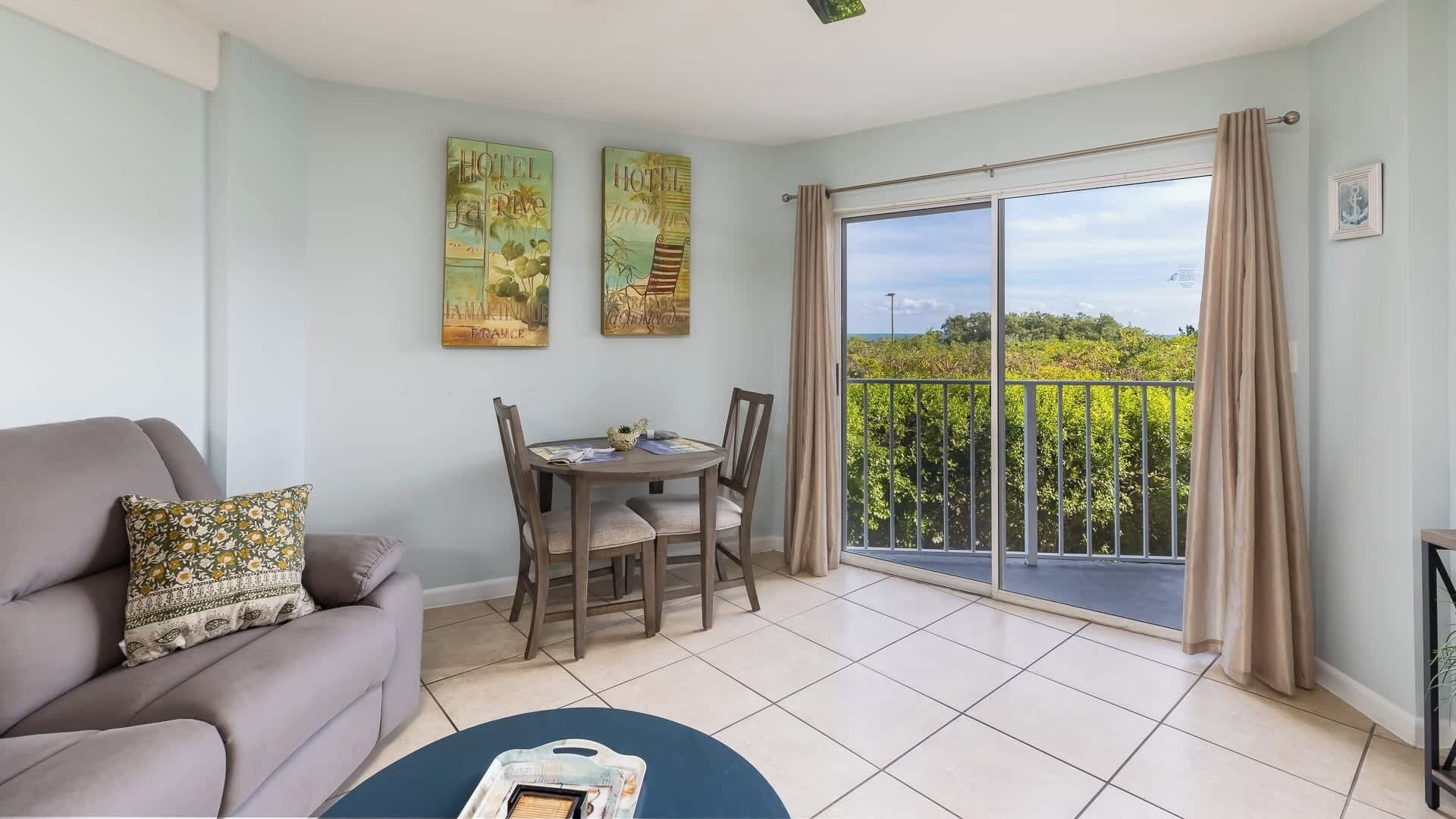 A room with a sofa, a dining table with chairs, a balcony with a view, and two wall art pieces.